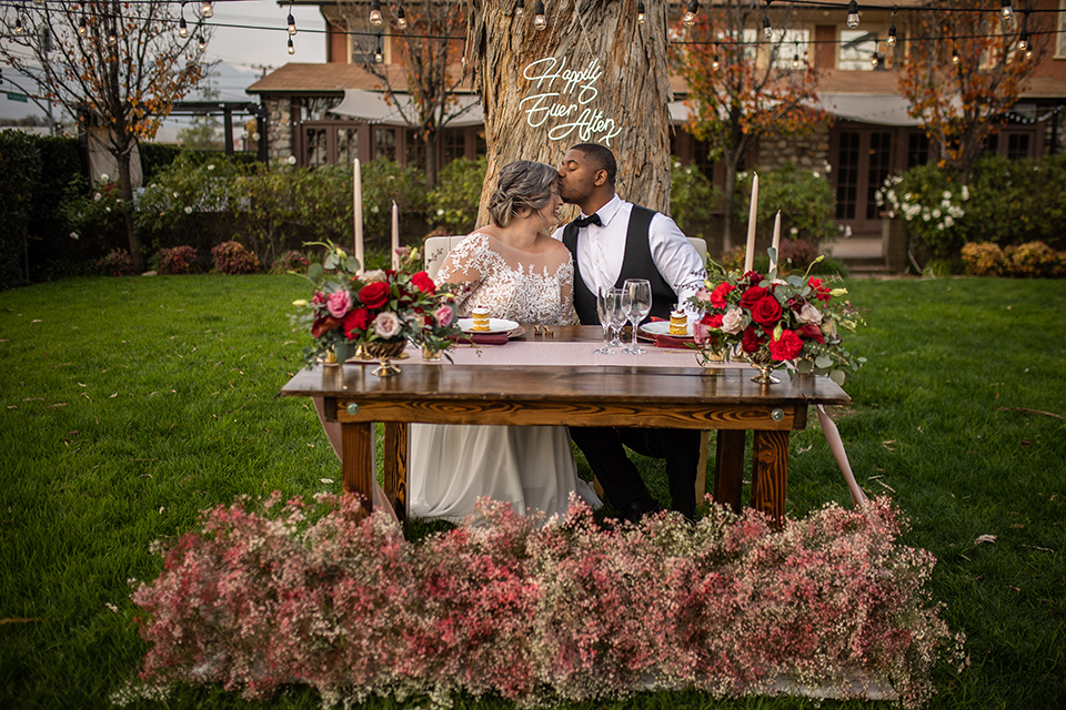  bride in a white and ivory gown with a natural waist and lace long sleeves, the groom in a burgundy velvet tuxedo with a black bow tie, and the bridesmaid in a red velvet long gown and a floral headpiece