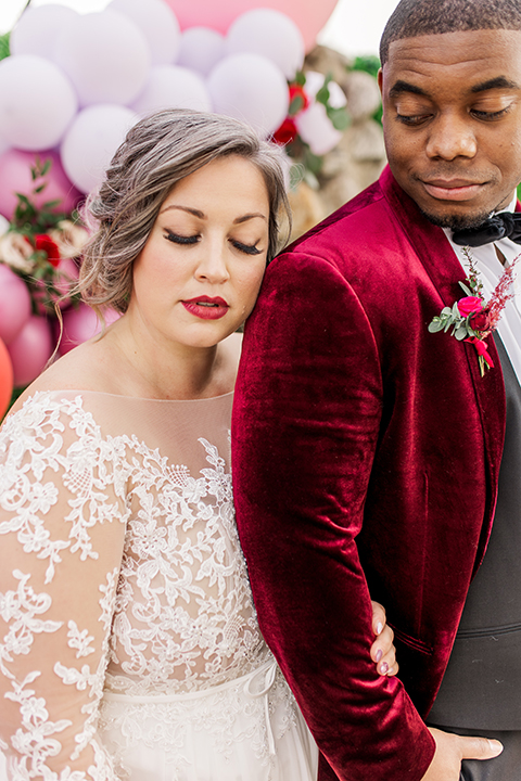  bride in a white and ivory gown with a natural waist and lace long sleeves, the groom in a burgundy velvet tuxedo with a black bow tie 