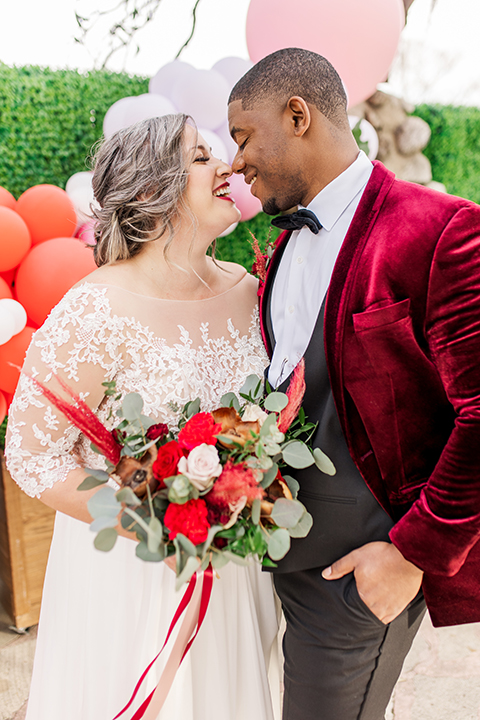  bride in a white and ivory gown with a natural waist and lace long sleeves, the groom in a burgundy velvet tuxedo with a black bow tie 