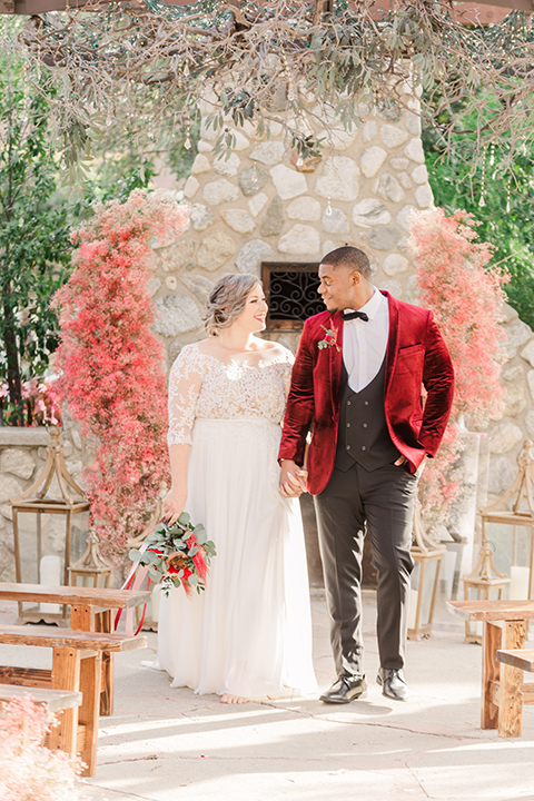  bride in a white and ivory gown with a natural waist and lace long sleeves, the groom in a burgundy velvet tuxedo with a black bow tie 