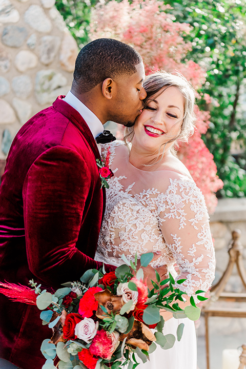  bride in a white and ivory gown with a natural waist and lace long sleeves, the groom in a burgundy velvet tuxedo with a black bow tie 