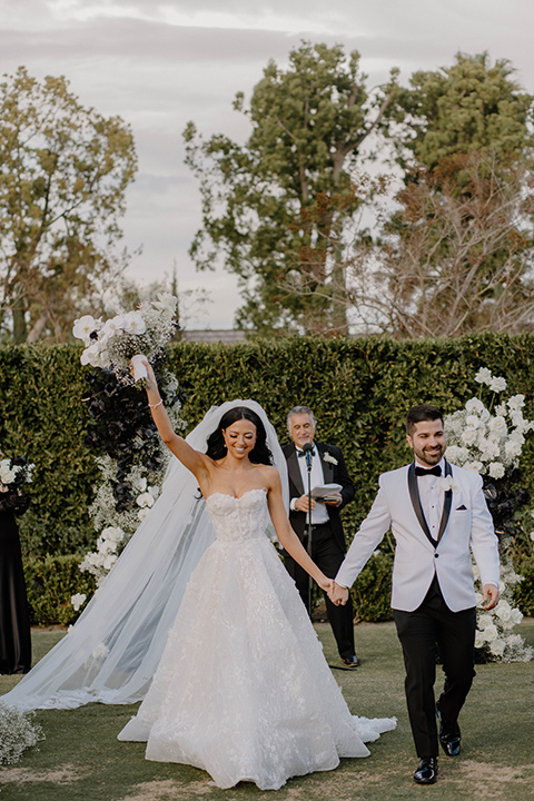  a chic black and white wedding with modern touches with the groom in a white tuxedo and the bride in a white ballgown and the bridesmaids and groomsmen in black dresses/tuxedos – walking down the aisle 