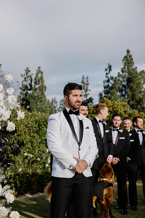  a chic black and white wedding with modern touches with the groom in a white tuxedo and the bride in a white ballgown and the bridesmaids and groomsmen in black dresses/tuxedos – groom seeing the bride walk down the aisle 