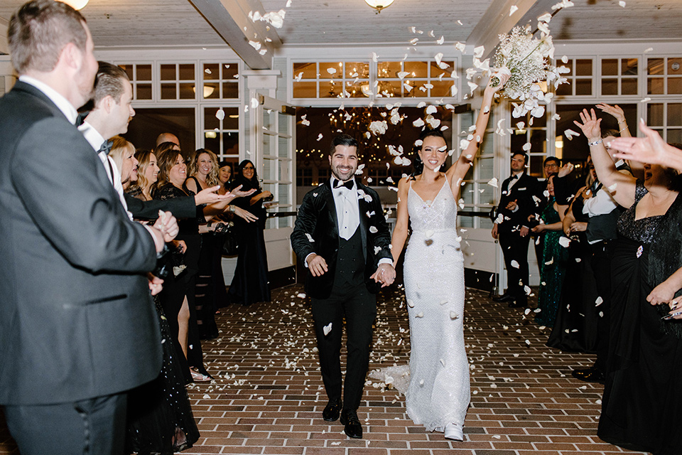  a chic black and white wedding with modern touches with the groom in a white tuxedo and the bride in a white ballgown and the bridesmaids and groomsmen in black dresses/tuxedos – couple leaving their wedding 