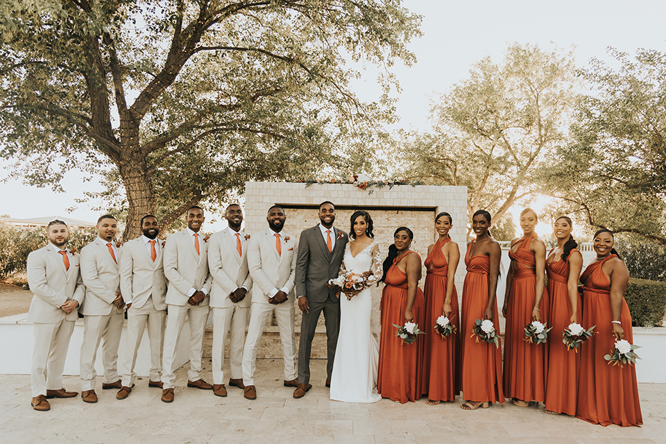 A dreamy amber toned wedding in Arizona at a wedgewood wedding venue – with the bride in a long sleeve lace gown and the bridesmaids in burnt orange dresses and the groom in a café brown suit and the groomsmen in a tan suit – picture of the bridal party in a line