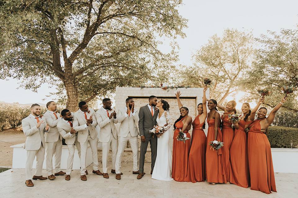 A dreamy amber toned wedding in Arizona at a wedgewood wedding venue – with the bride in a long sleeve lace gown and the bridesmaids in burnt orange dresses and the groom in a café brown suit and the groomsmen in a tan suit – picture of the bridal party in a line