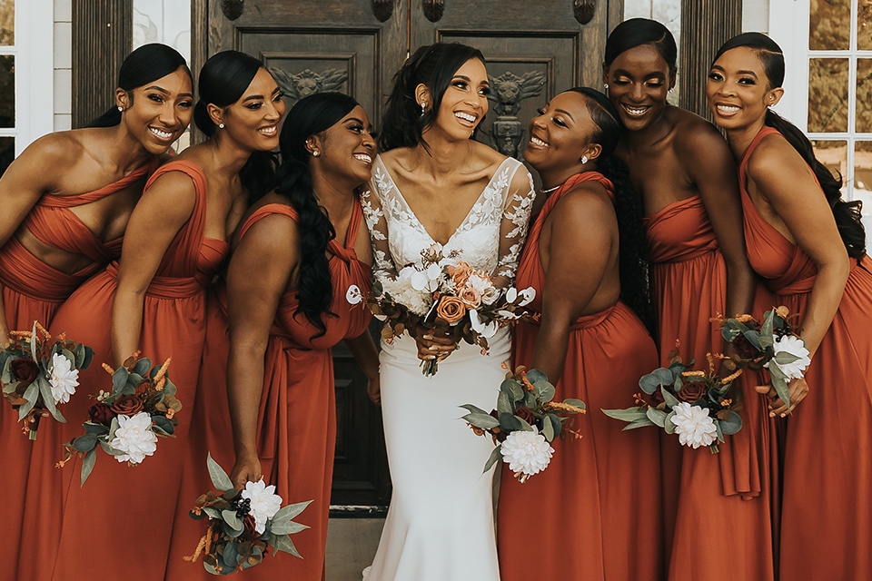 A dreamy amber toned wedding in Arizona at a wedgewood wedding venue – with the bride in a long sleeve lace gown and the bridesmaids in burnt orange dresses and the groom in a café brown suit and the groomsmen in a tan suit – picture of the bridesmaids hugging the bride