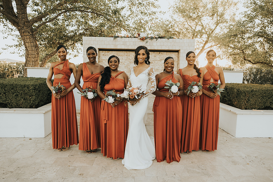 A dreamy amber toned wedding in Arizona at a wedgewood wedding venue – with the bride in a long sleeve lace gown and the bridesmaids in burnt orange dresses and the groom in a café brown suit and the groomsmen in a tan suit – picture of the in a line