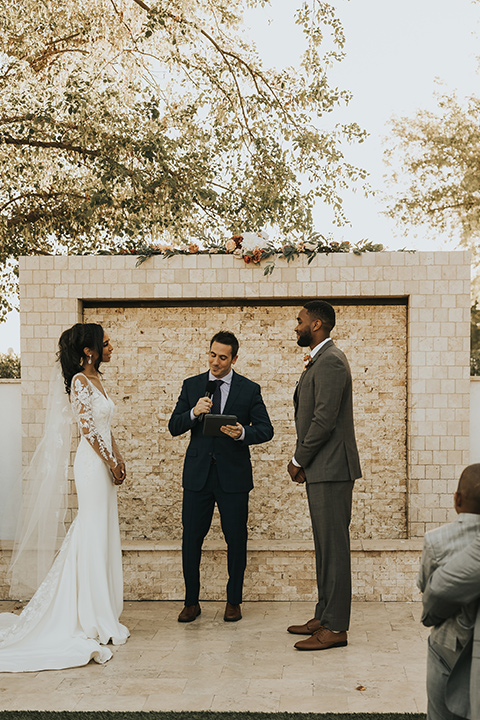  A dreamy amber toned wedding in Arizona at a wedgewood wedding venue – with the bride in a long sleeve lace gown and the bridesmaids in burnt orange dresses and the groom in a café brown suit and the groomsmen in a tan suit – couple at the ceremony 