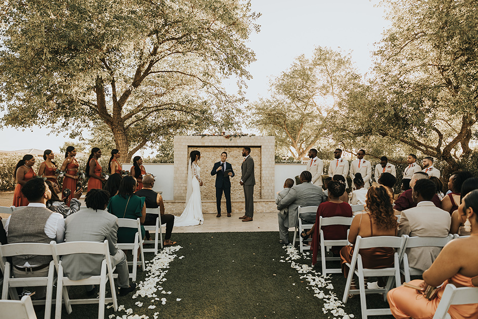 A dreamy amber toned wedding in Arizona at a wedgewood wedding venue – with the bride in a long sleeve lace gown and the bridesmaids in burnt orange dresses and the groom in a café brown suit and the groomsmen in a tan suit – couple at the ceremony