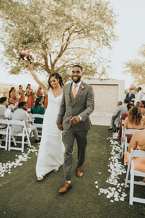  A dreamy amber toned wedding in Arizona at a wedgewood wedding venue – with the bride in a long sleeve lace gown and the bridesmaids in burnt orange dresses and the groom in a café brown suit and the groomsmen in a tan suit – couple at the ceremony walking down the aisle