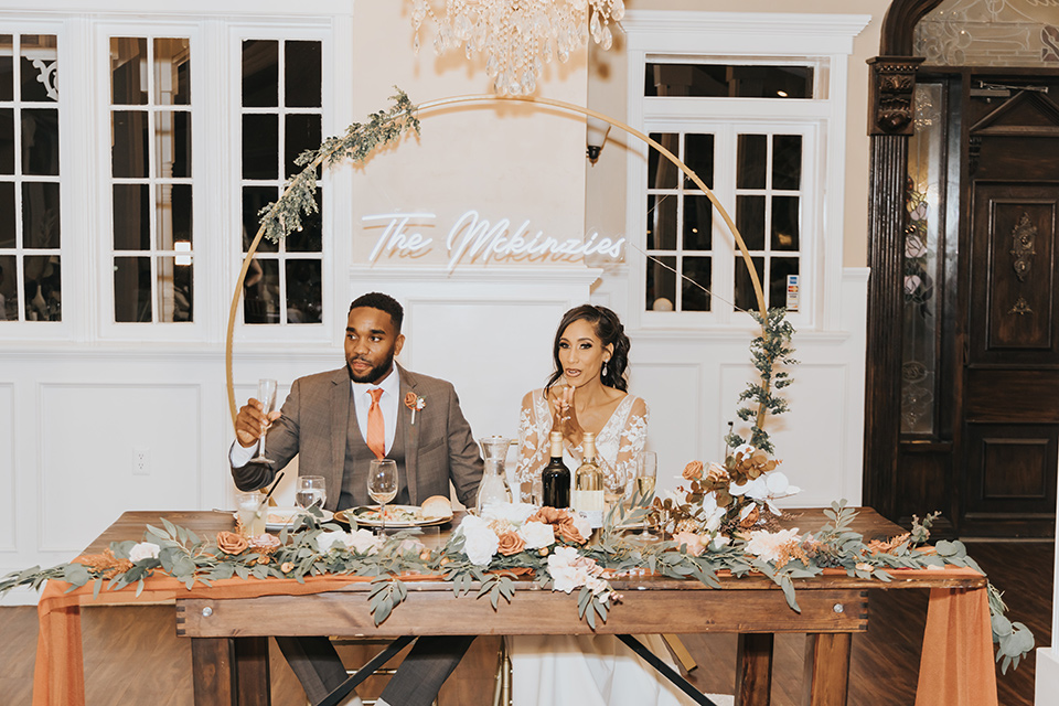 A dreamy amber toned wedding in Arizona at a wedgewood wedding venue – with the bride in a long sleeve lace gown and the bridesmaids in burnt orange dresses and the groom in a café brown suit and the groomsmen in a tan suit – couple sitting at the sweetheart table together
