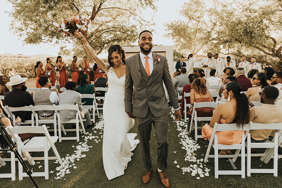 A dreamy amber toned wedding in Arizona at a wedgewood wedding venue – with the bride in a long sleeve lace gown and the bridesmaids in burnt orange dresses and the groom in a café brown suit and the groomsmen in a tan suit – couple walking down the aisle after the ceremony