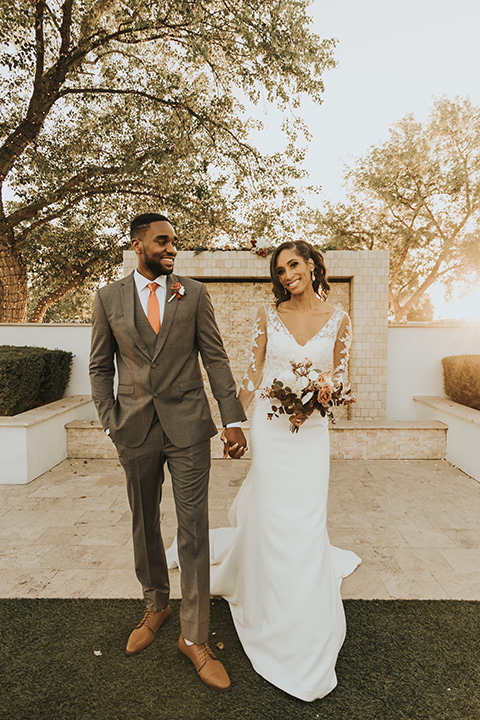  A dreamy amber toned wedding in Arizona at a wedgewood wedding venue – with the bride in a long sleeve lace gown and the bridesmaids in burnt orange dresses and the groom in a café brown suit and the groomsmen in a tan suit – couple walking toward camera 