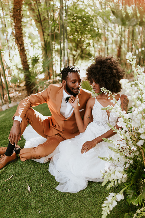  bohemian wedding with the bride in a flowing lace gown and the groom in a caramel brown suit with a green velvet bow tie- couple sitting in the grass together 