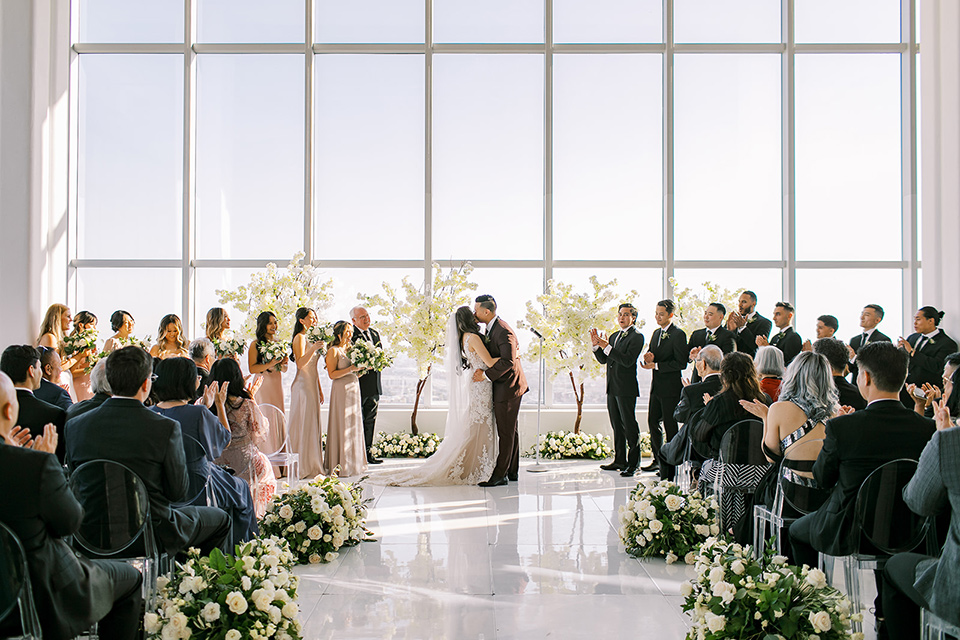 A city wedding with the guys in burgundy and black tuxedos and the bridesmaids in blush dresses- first kiss at ceremony