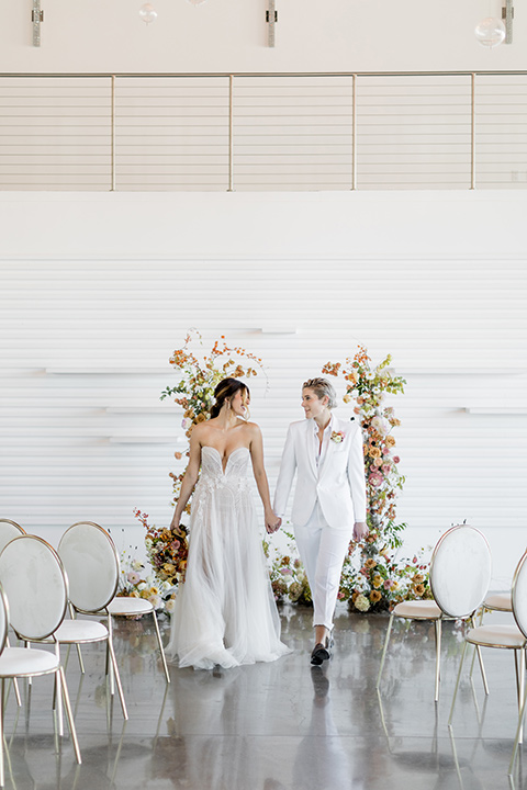 Modern romantic city wedding with an all-white color scheme and bright florals – one bride wore a strapless a-line gown and the other bride wore a white suit – brides walking down the aisle 