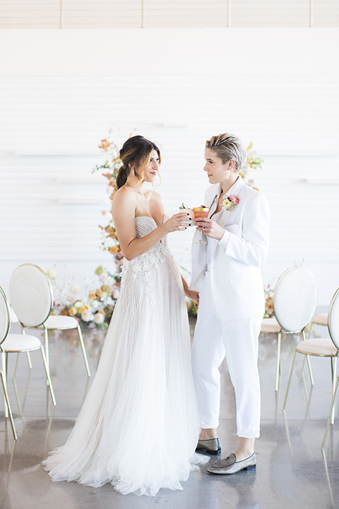 Modern romantic city wedding with an all-white color scheme and bright florals – one bride wore a strapless a-line gown and the other bride wore a white suit – brides cheersing