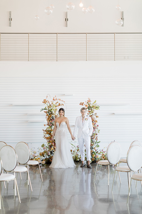 Modern romantic city wedding with an all-white color scheme and bright florals – one bride wore a strapless a-line gown and the other bride wore a white suit – brides walking down the aisle