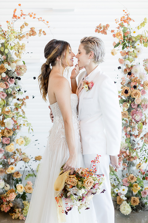 Modern romantic city wedding with an all-white color scheme and bright florals – one bride wore a strapless a-line gown and the other bride wore a white suit – brides heads close 