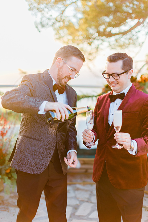  romantic winter inspired wedding theme with luxe textures and details, one groom wore a burgundy velvet shawl tuxedo and the other groom in a black paisley tuxedo – grooms cheersing with champagne