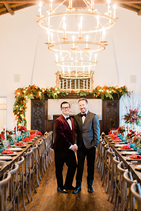  romantic winter inspired wedding theme with luxe textures and details, one groom wore a burgundy velvet shawl tuxedo and the other groom in a black paisley tuxedo – grooms walking into their reception