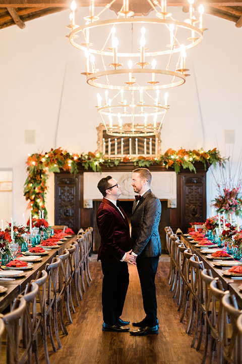  romantic winter inspired wedding theme with luxe textures and details, one groom wore a burgundy velvet shawl tuxedo and the other groom in a black paisley tuxedo – grooms walking into their reception 