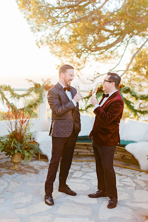  romantic winter inspired wedding theme with luxe textures and details, one groom wore a burgundy velvet shawl tuxedo and the other groom in a black paisley tuxedo – grooms cheersing with champagne 