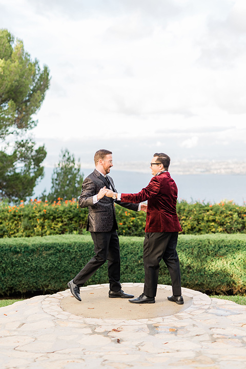  romantic winter inspired wedding theme with luxe textures and details, one groom wore a burgundy velvet shawl tuxedo and the other groom in a black paisley tuxedo – grooms dancing outside at the venue