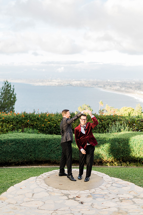  romantic winter inspired wedding theme with luxe textures and details, one groom wore a burgundy velvet shawl tuxedo and the other groom in a black paisley tuxedo – grooms dancing outside at the venue 