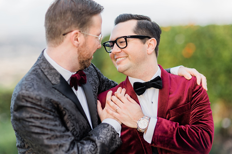  romantic winter inspired wedding theme with luxe textures and details, one groom wore a burgundy velvet shawl tuxedo and the other groom in a black paisley tuxedo – grooms embracing