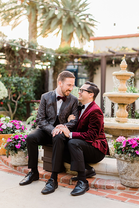  romantic winter inspired wedding theme with luxe textures and details, one groom wore a burgundy velvet shawl tuxedo and the other groom in a black paisley tuxedo – grooms walking in the garden