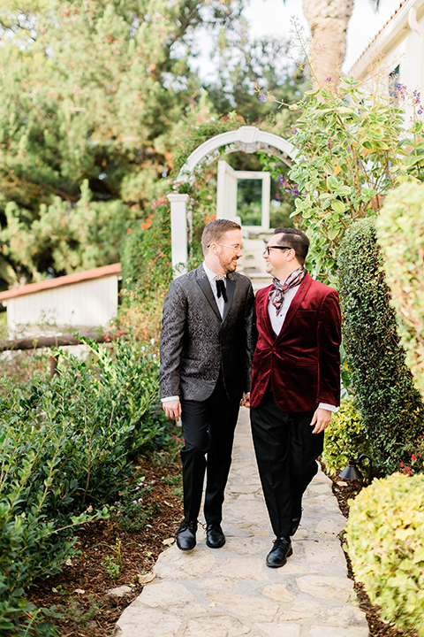  romantic winter inspired wedding theme with luxe textures and details, one groom wore a burgundy velvet shawl tuxedo and the other groom in a black paisley tuxedo – grooms walking in the garden