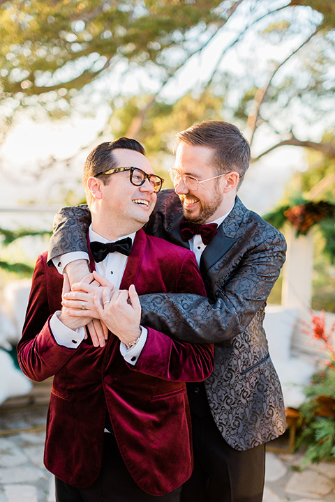  romantic winter inspired wedding theme with luxe textures and details, one groom wore a burgundy velvet shawl tuxedo and the other groom in a black paisley tuxedo – grooms sitting by the fountain 