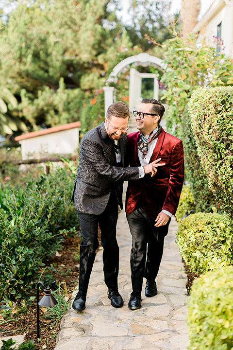  romantic winter inspired wedding theme with luxe textures and details, one groom wore a burgundy velvet shawl tuxedo and the other groom in a black paisley tuxedo – grooms walking in the garden 