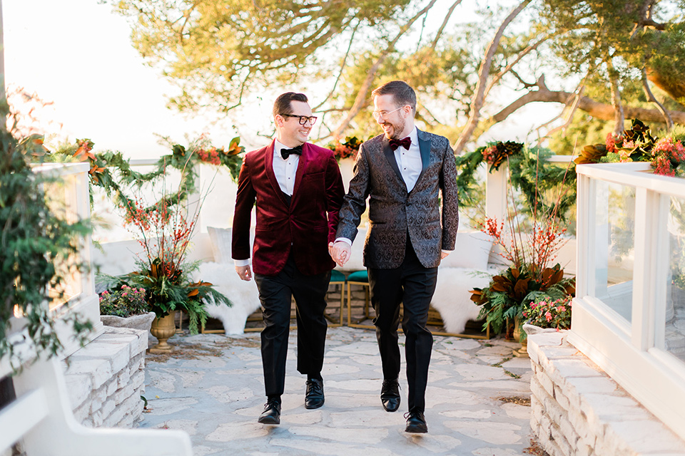  romantic winter inspired wedding theme with luxe textures and details, one groom wore a burgundy velvet shawl tuxedo and the other groom in a black paisley tuxedo – the grooms walking and holding hands