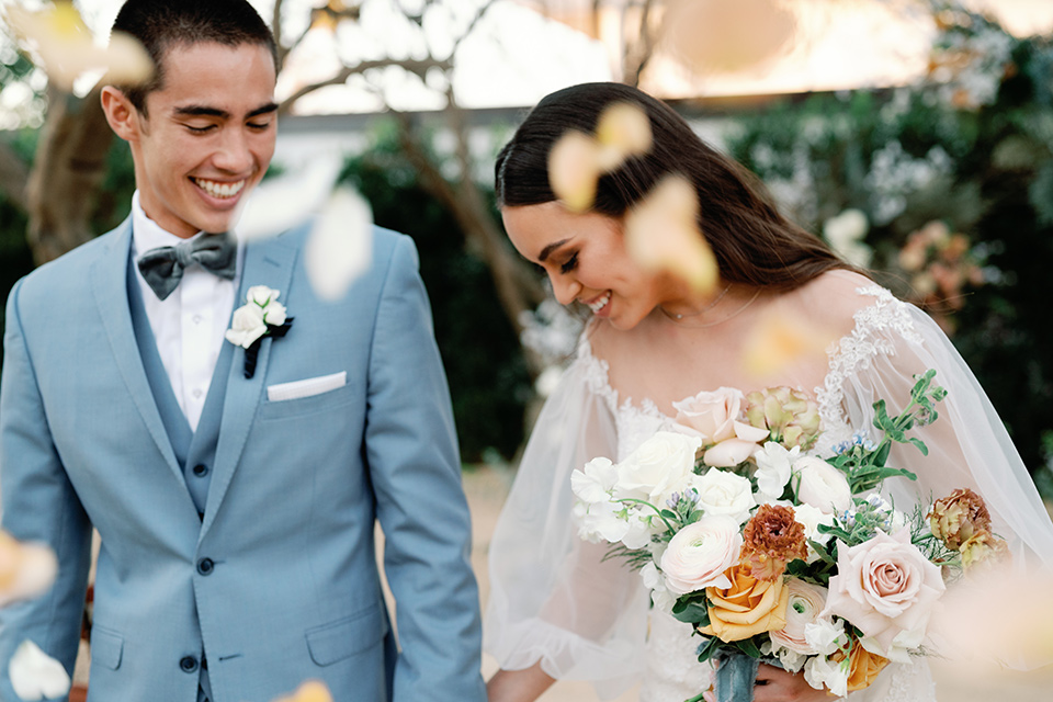  a wedding at the grand gimeno with white, blue, and gold accents – the bride wore a long sleeved modern fitted gown and the groom wore a light blue suit with velvet bow tie – bride and groom walking with confetti 