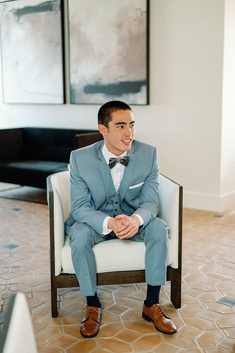 a wedding at the grand gimeno with white, blue, and gold accents – the bride wore a long sleeved modern fitted gown and the groom wore a light blue suit with velvet bow tie – groom sitting in the chair 