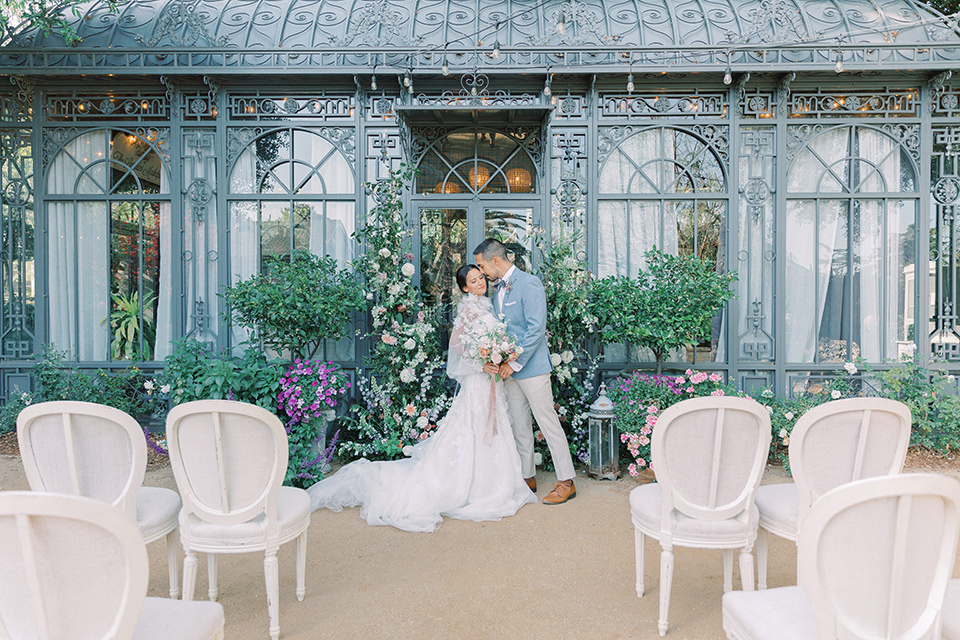 A French inspired wedding at a lavender farm with the bride in a fashion-forward white a-line gown with a high neckline and three dimensional floral appliques on it, the groom wore a light blue suit coat with a pair of tan pant and a grey velvet bow tie, they are kissing at the ceremony