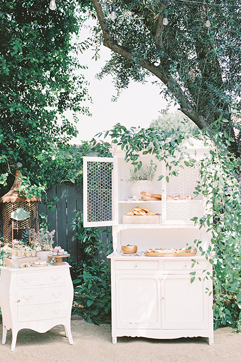  A French inspired wedding at a lavender farm with the bride in a fashion-forward white a-line gown with a high neckline and three dimensional floral appliques on it, the groom wore a light blue suit coat with a pair of tan pant and a grey velvet bow tie, the décor has fun antique pieces and this image shows a white cabinet with cocktails and charcutarie on it
