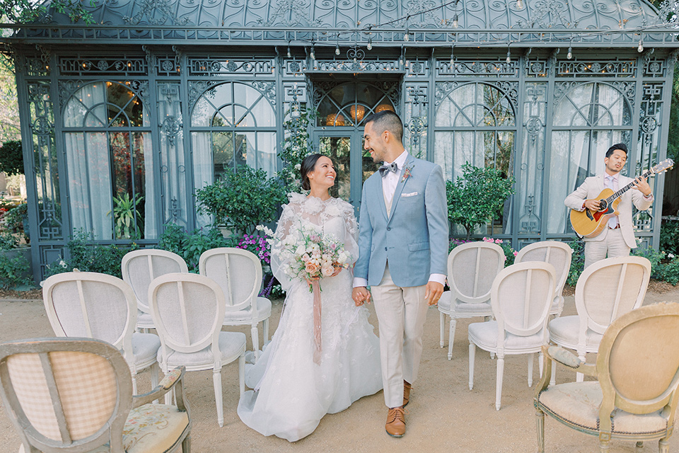 A French inspired wedding at a lavender farm with the bride in a fashion-forward white a-line gown with a high neckline and three dimensional floral appliques on it, the groom wore a light blue suit coat with a pair of tan pant and a grey velvet bow tie, they are walking down the aisle after the ceremony