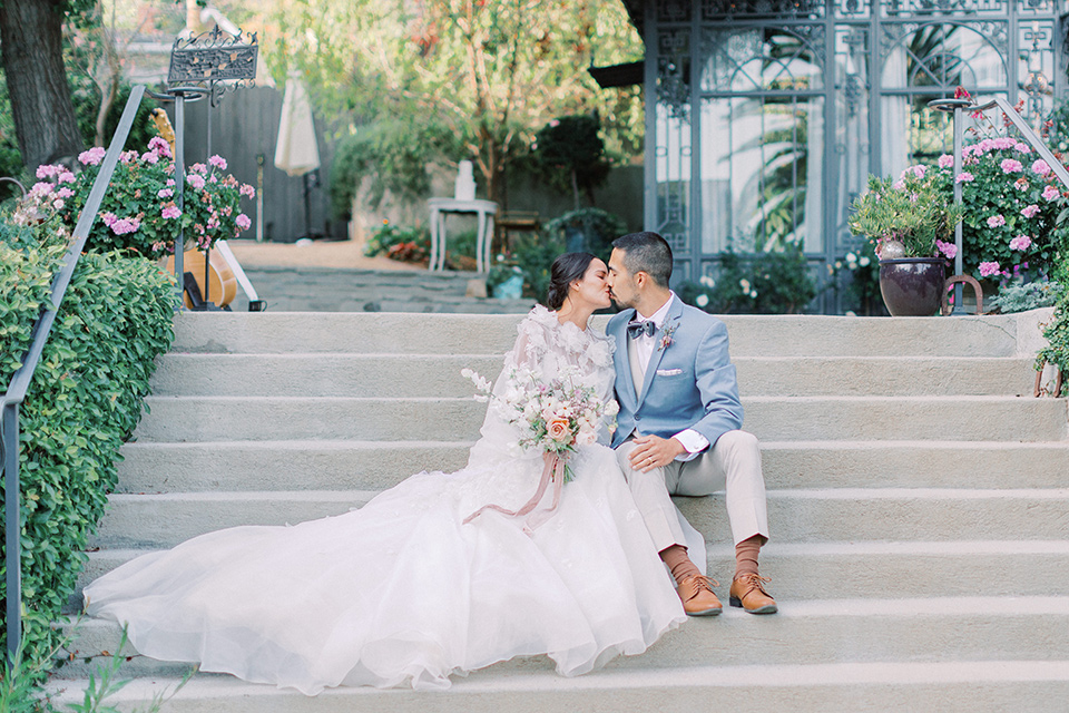 A French inspired wedding at a lavender farm with the bride in a fashion-forward white a-line gown with a high neckline and three dimensional floral appliques on it, the groom wore a light blue suit coat with a pair of tan pant and a grey velvet bow tie, they are sitting and kissing on the steps overlooking the garden