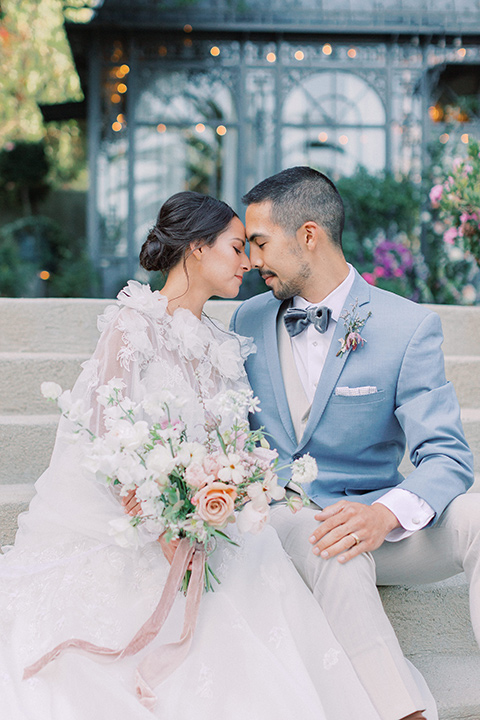  A French inspired wedding at a lavender farm with the bride in a fashion-forward white a-line gown with a high neckline and three dimensional floral appliques on it the groom wore a light blue suit coat with a pair of tan pant and a grey velvet bow tie, couple at the ceremony 