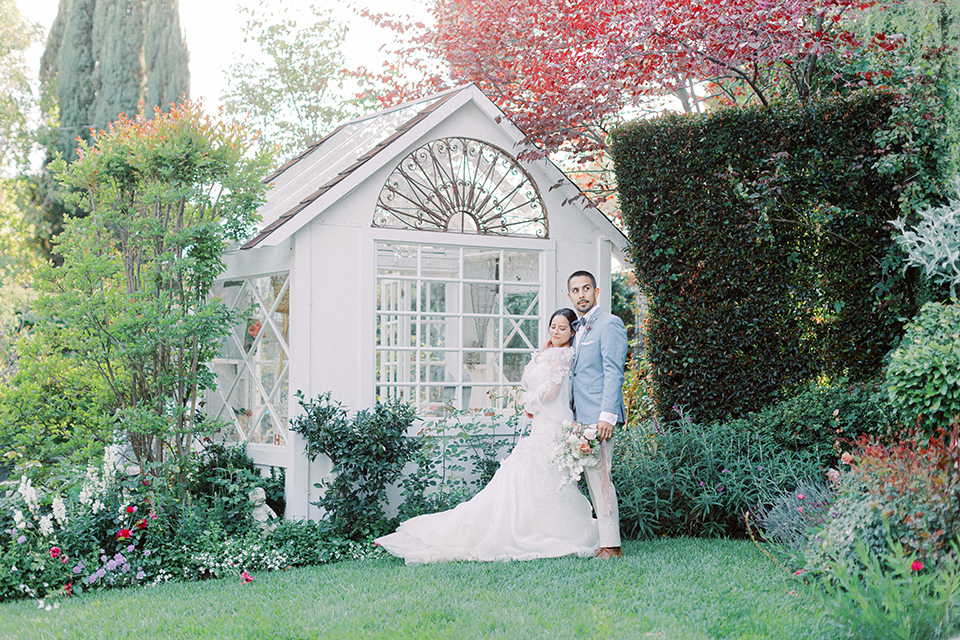 A French inspired wedding at a lavender farm with the bride in a fashion-forward white a-line gown with a high neckline and three dimensional floral appliques on it, the groom wore a light blue suit coat with a pair of tan pant and a grey velvet bow tie, they are dancing in the garden