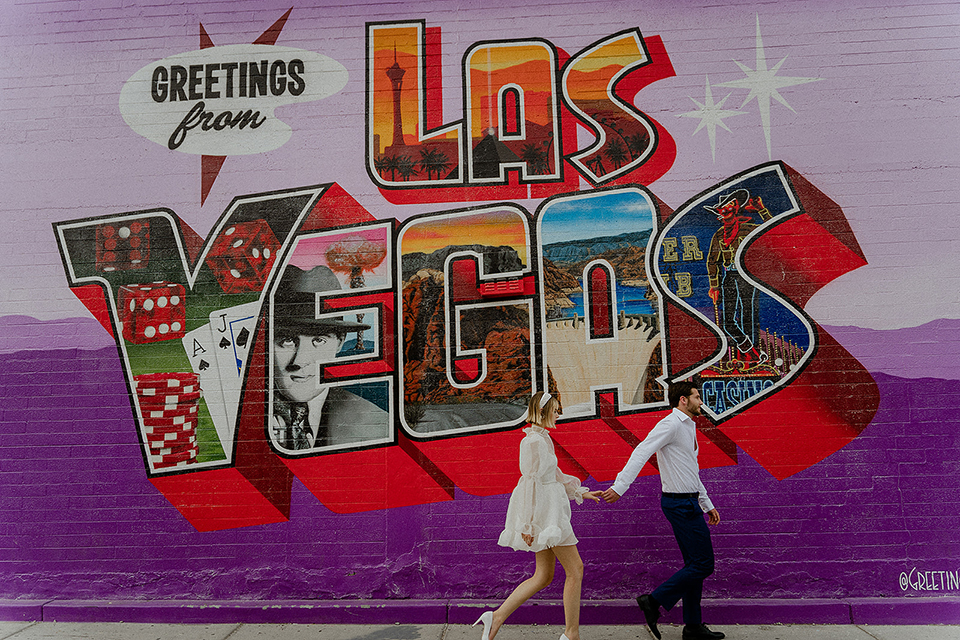  how to rent a suit or tuxedo in vegas young couple walking hand in hand down the sidewalk in las vegas with a las vegas sign painted on a brick wall with a purple background
