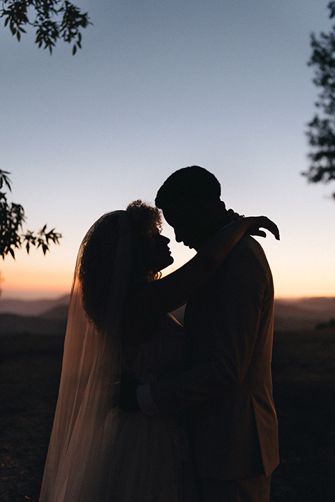  an elevated cottagecore wedding design in a barn venue with gold and tan tones – the bride in a white gown with long sleeves and the groom in a tan suit – couple at sunset 