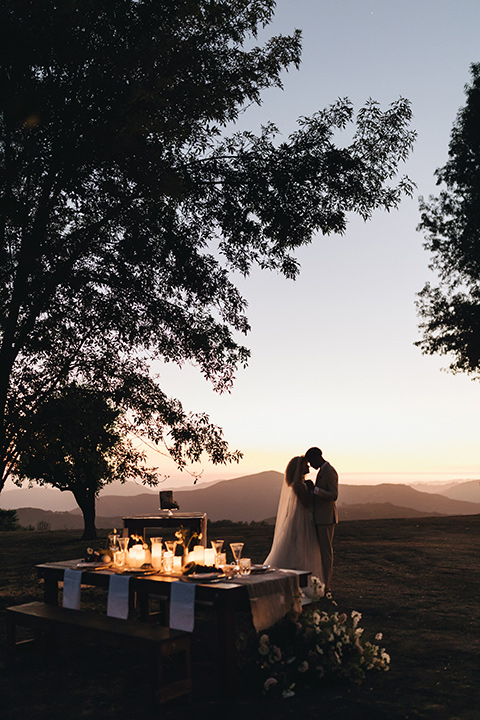  an elevated cottagecore wedding design in a barn venue with gold and tan tones – the bride in a white gown with long sleeves and the groom in a tan suit – couple at sunset 