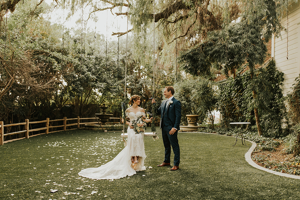  a romantic rustic garden wedding with the groom in a blue suit and floral tie and the bride in a lace gown - couple by the swings 