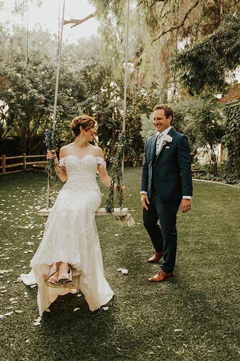  a romantic rustic garden wedding with the groom in a blue suit and floral tie and the bride in a lace gown - groom pushing bride on the swing 