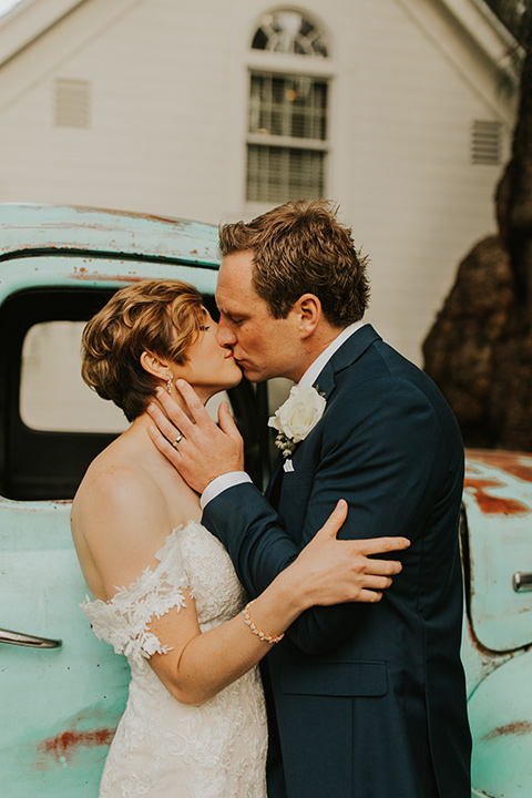  a romantic rustic garden wedding with the groom in a blue suit and floral tie and the bride in a lace gown - couple kissing in front of rustic truck 